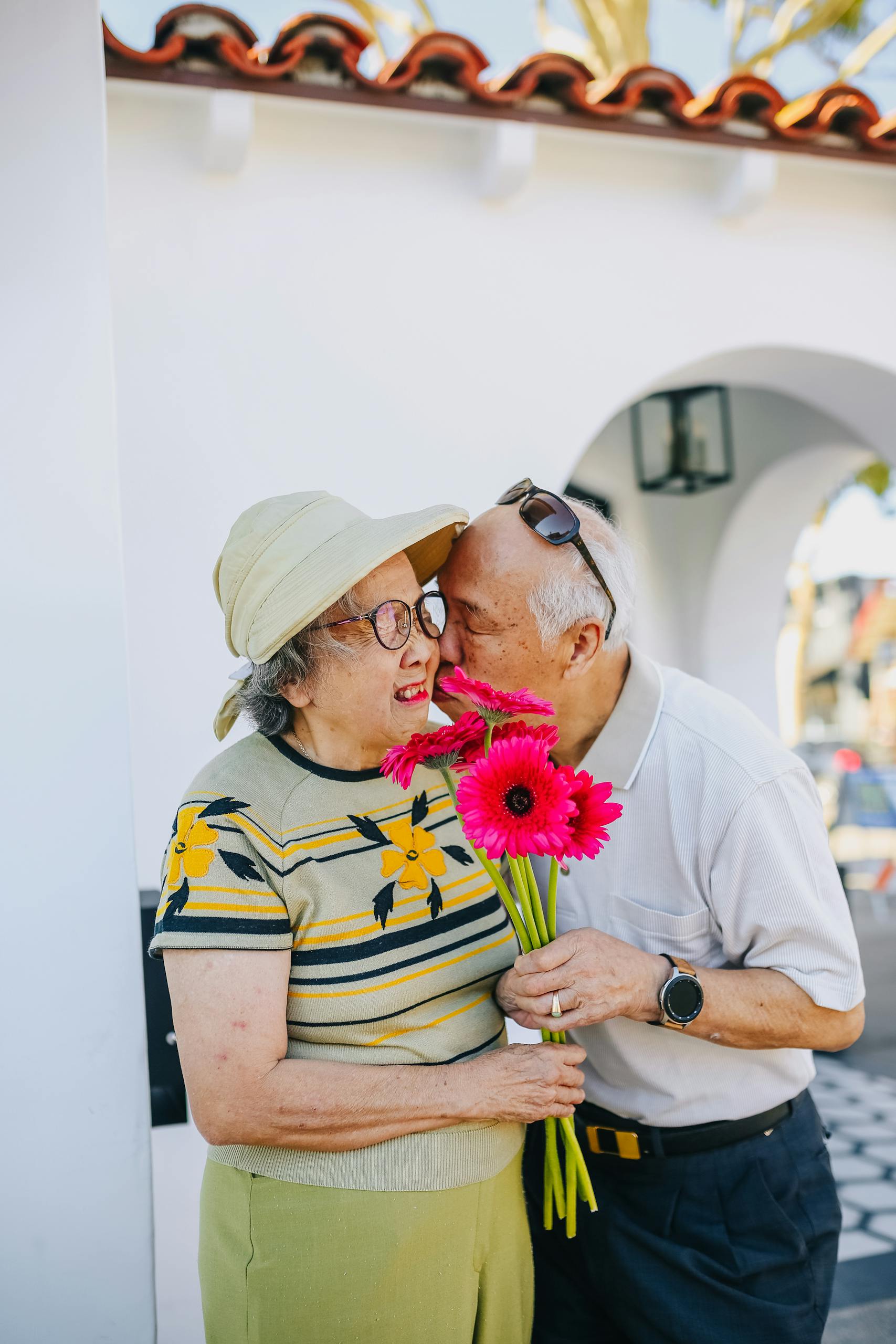 A loving elderly couple shares a tender moment outdoors, holding vibrant pink flowers.